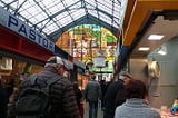 Shoppers in an indoor open-air market with a glass roof and stained-glass windows.