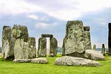 Color photo closeup of at least eight stones from UK’s Stonehenge during a semi-cloudy mixed with little bits of pale blue sky daylight, amidst verdant green grass. The focal center point is a classic two large stones with a cross stone atop them, as if a natural archway one my see and pass through.