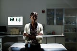 A photo of a Black female doctor sitting in an examination room, arms crossed.