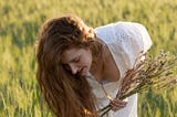 Red-haried woman with long hair bends down to gather a bunch of long blooming grass on a very sunny day. She is dressed in an airy white dress. You can see down her blouse a bit, but the picture is unprovocative. You see long grass all around here.