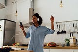 A photo of a black woman listening to music on headphones in her kitchen. She is feeling herself.