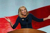 Jill Biden speaking on stage in front of a large LCD screen with a wavy red stripe on a white background.