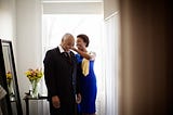 A Black woman assists her elder father with his collar as they prepare for a party.