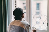 Black woman holding a cup of coffee while looking out her window.