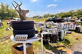 Color horizontal photo of a low-cut blue sky sunny day grassy field covered in folding tables, a distant open-bed black truck, tons of local farmers market goods all over. In the foreground as partial closeup left to right in the frame we see featured a white round table with 2 dozen-egg cartons, 2 fat black tires with a deer head in profile to us sticking out from the top one, and below the table a box of items beneath it, plus a drumkit on the grass to the right of this table, etc. around.