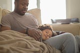 A photo of a young black girl sleeping with her head on her dad’s lap.