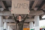 A woman stands beneath an overpass. With her hands, she’s holding high a cardboard sign with the word WOMEN on it.