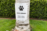 A sign saying “dog library” on a tree with a pile of sticks in front of it