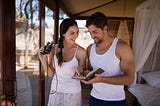 A couple in their bedroom, smiling. A black-haired woman is holding binoculars while a bearded man in a white shirt is pointing to something in a book he is holding open.