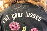 Color photo showing the back of the hair stylist’s black t-shirt, upon which is the wording “cut your losses” with a graphic image of a skeleton hand holding a pair of scissors that are snipping 2 pink roses from their stems.