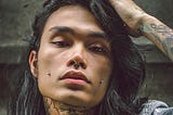 A man of Asian ethnicity poses in a close-up shot with his left hand resting on his long, dark hair. He has a nose piercing, and two cheeks pierced with silver studs. He also has tattoos on his neck and left forearm. He’s wearing a denim jacket.