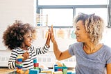 A photo of a mom and her young child high fiving as the kid plays with blocks.