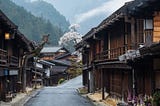 A scenic view of Tsumago-juku, a historic post town on the Nakasendo, during spring. Traditional Edo period (1603–1868) wooden buildings line the quiet street, with a cherry blossom tree in full bloom in the background. Misty mountains and lush greenery enhance the nostalgic atmosphere of this well-preserved town.