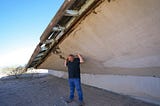 A person in black shirt and jeans standing under the foundation of a wooden house, seemingly keeping it from collapsing with his bare hands.