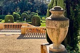 A labyrinth, as seen from above. There is a statue in the middle of the maze. There are tall trees in the background, and we are viewing the scene from a vantage point of some steps in a stately courtyard.