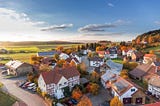 A high-angle view of a town with trees turning color and a few clouds in a blue sky.