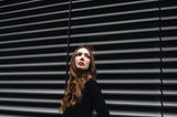 A woman wearing a chic black outfit is leaning against a gray, shuttered wall.