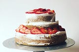 A layered cake on a silver cake stand — the cake is golden brown with lightly applied white icing and strawberries.