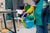 A waist-down shot of a maid, seen from behind, holding a bucket of cleaning products in a small room.