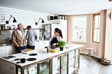 A photo of an Asian woman in the kitchen with her white husband and father-in-law.