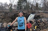 Photo of woman standing with two bicycles in front of a rock retaining wall with a waterfall in the background. Alt text to demonstrate the importance of alt text.