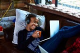 Smiling Black woman with headphones on looking at her computer with a cup of coffee in her hand.