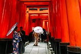 The overwhelmingly red view of Fushimi Inari Shrine in Kyoto, Japan. Two woman are dressed in traditional kimonos with outfit matching umbrellas. A man, also in traditional garb, is taking a photo of one of the women.