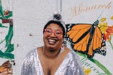 Sondra — a Black woman — poses in a sparkling silver gown. Behind her is a mural showing butterflies in bright colors.