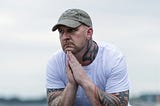 Man sitting outside on a rock looking lost in thought and a bit sad. Pensive. He is wearing a white t-shirt and jeans, with a green ballcap. His arms are tattooed. He has workboots on. The background is blurred but looks like a beach’s rocky shore.