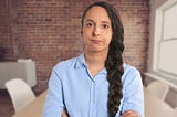 Woman with long braided hair and a blue shirt standing inside a red-brick room with her arms crossed looking annoyed