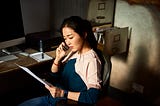 A woman looks serious as she listens on the phone while holding paperwork and sitting in an office chair.