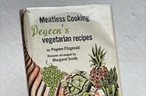 A hand holding up a vintage cookbook with a quaint illustration of fruits and veggies on the front.