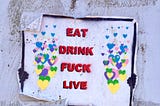 Street art graffiti stencil of a black-booted person holding up a large sign, covering the rest of their body and face, with colorful hearts bursting on either side of the message stating in all-red-capital letters “EAT DRINK FUCK LIVE”.