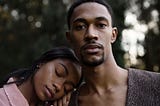 A photo of a black woman resting her head on the shoulder of a black man who is looking at the camera.