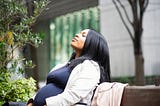 A photo of a pregnant black woman sitting outside on a bench, closing her eyes with a contemplative expression.