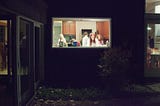 Family group washing dishes in the kitchen after a Thanksgiving meal, shot from outdoors through a picture window at night.