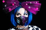 A person with a blue bob haircut wearing a huge purple hairbow and a mask with shiny bats + moons and purple spiderwebs
