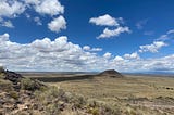 A mostly flat New Mexican landscape with scrubby grass under a deep blue sky with white fluffy clouds.
