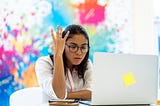 A woman throws her arm up in frustration as she stares at her laptop.