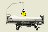 Side-view of a hospital bed with a metal bar above it; hanging from it is a triangular yellow caution sign.