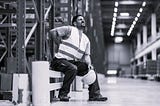 Man sitting in warehouse with a painful grimace on his face while holding his back. He is wearing a work vest and holding a hard hat.
