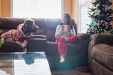 A smiling, long-haired person wearing pajamas and holding a mug sitting on the couch next to a dog and a Christmas tree.