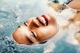 “Embodiment and Reconnecting to Self: Embrace Inner Harmony” This photo shows the face of a woman with carmel-brown skin floating in a pool of blue water. Only her face is above the water and she is smiling.