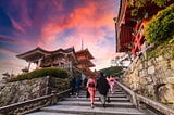 Visitors in kimono climbing the stone steps to Kyoto’s Kiyomizu-dera at sunset, with its pagoda and temple gates in view.
