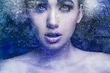 A woman with light blue eyes and purple lipstick poses with a shocked expression, the cool tones and frosted effects of the shot indicate she’s encased beneath a sheet of ice.