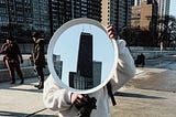 Person holding a camera in one hand and a mirror with a highrise building inside in the other. They’re wearing grey jeans, a white fluffy jacket, and white Nike sneakers.