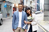A photo of a happy Asian couple walking down the street. The woman is holding a bouquet of white flowers.
