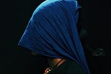 a person in profile view against a black background. They are wearing a vibrant blue headscarf or veil that completely covers their head and face. The person is dressed in a dark green top or sweater and has a colorful beaded necklace visible at their neck, featuring orange, red, and multi-colored beads. The composition is striking in its contrast between the deep blue of the head covering and the darkness of the background, creating a contemplative, mysterious atmosphere.