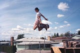 A teenage boy in a t-shirt, shorts, and a baseball cap holds his skateboard to his feet after launching himself in the air from a plywood ramp, framed against the sky above the homes of a working-class neighborhood in 1980s Spokane.