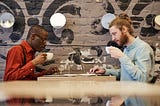 A black man and a white man in front of their laptops sipping coffee directly across from each other at a cafe table.